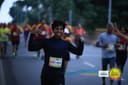 A runner in a navy quarter-zip flexing both arms mid-race at the Bengaluru Runners Jatre, holding a phone and a tube of electrolytes