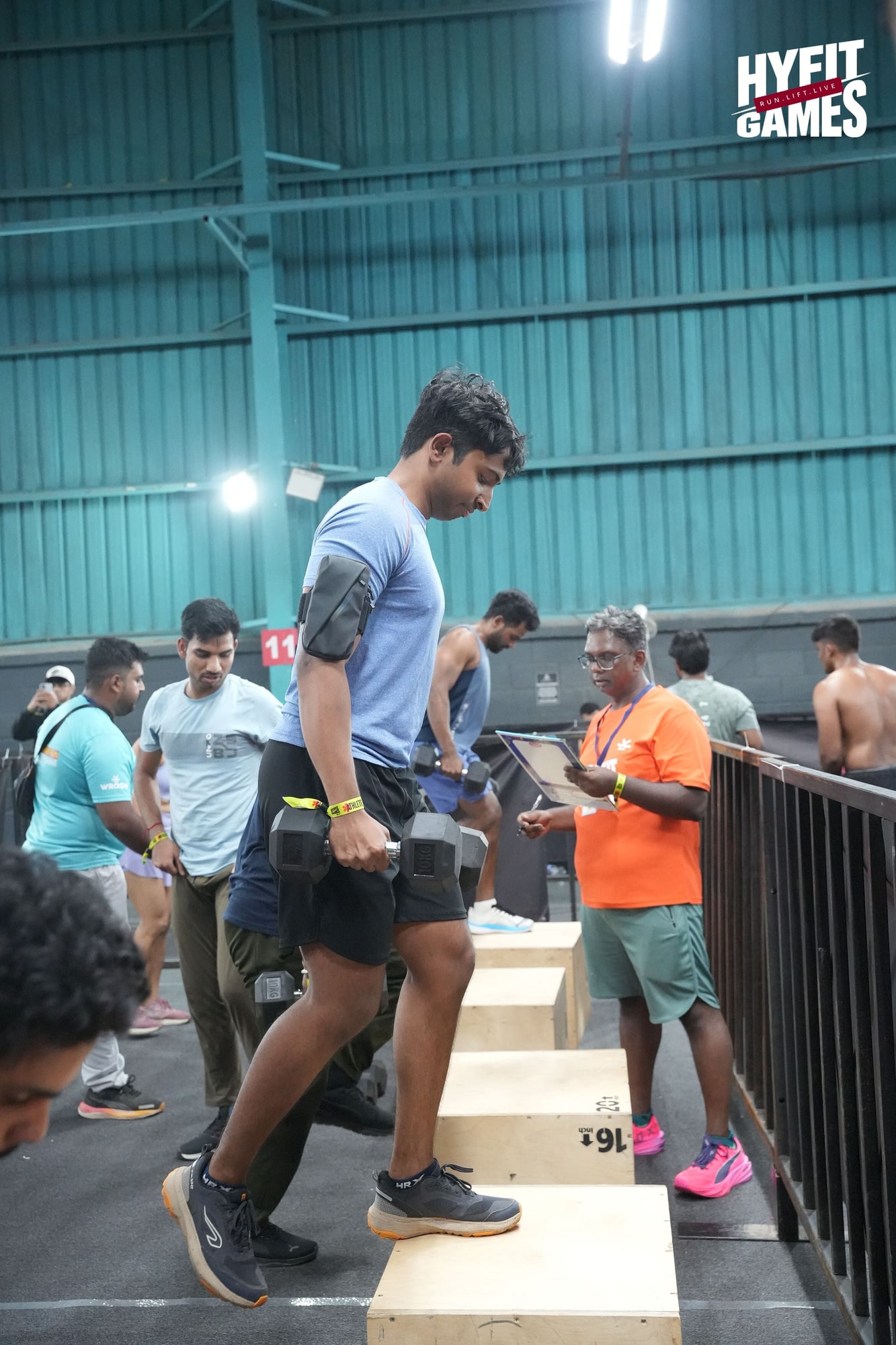 An athlete carrying two dumbbells up a wooden box at HYFIT Games Bengaluru, a judge counting reps beside him